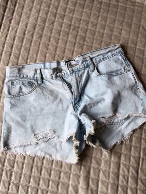 Ljusblå denimshorts med fransig kant - Säljer ett par ljusblå denimshorts med fransig kant och slitna detaljer framtill. Klassisk femficksmodell med knapp och dragkedja. Perfekta för sommaren och ger en avslappnad vibe till din outfit.