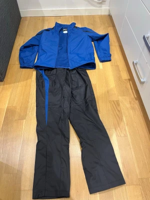 Blå och svart träningsdress från south west - Säljer en sportig träningsdress med blå jacka och svarta byxor med blåa detaljer längs sidorna. Jackan har dragkedja och resår vid ärmsluten, byxorna har resår i midjan och dragsko. Perfekt för träning eller uppvärmning. Materialet är troligtvis polyester.