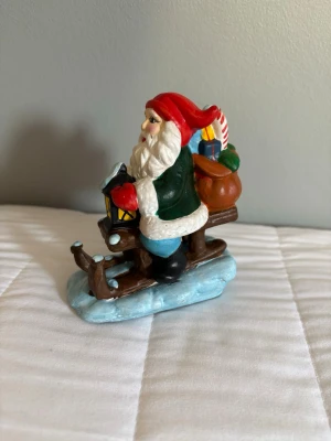 Vintage Tomtefigur på kälke med paket - ca 11x15 cm,Dekorativ tomtefigur i färgglad design, sittande på en brun kälke med blått underlag. Tomten har röd luva, grön jacka, blå byxor och håller en lykta. Bakom honom finns en säck med paket och en stor polkagris. Perfekt som juldekoration.