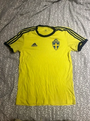 Gul Sverige fotbollströja Adidas - Gul kortärmad fotbollströja från Adidas med svarta detaljer och Sveriges landslagsemblem på bröstet. Svarta ränder på axlarna och svart krage. Perfekt för match eller träning. Materialet är lätt och andas bra.