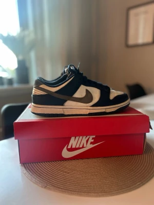 Nike Dunk Low svart/vit sneaker - Säljer ett par Nike Dunk Low sneakers i svart och vitt. Skorna har klassisk låg siluett, vita paneler i läder och svarta overlays samt Nike swoosh på sidan. Perfekta för dig som gillar streetwear och vill ha en ikonisk sneaker i garderoben.