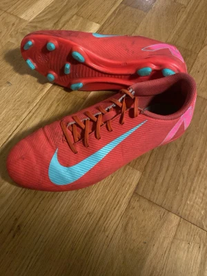 Röda Nike fotbollsskor med turkosa detaljer - Säljer ett par röda Nike fotbollsskor med turkosa detaljer och dobbar. Skorna har snörning och en låg profil för bra bollkontroll. Swoosh-loggan syns tydligt på sidan och hälen har rosa mönster. Perfekta för dig som vill sticka ut på planen.