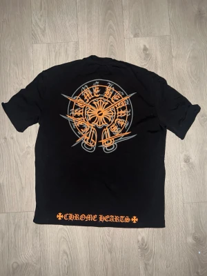 Svart Chrome Hearts t-shirt med tryck - Säljer en svart t-shirt från Chrome Hearts med stort orange och grått tryck på ryggen och mindre tryck på bröstet. Nedtill på ryggen står det 'Chrome Hearts' i orange. Klassisk passform lite baggy och rund hals, perfekt för dig som gillar streetwear. 