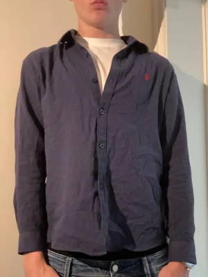Mörkblå skjorta från Polo Ralph Lauren - Klassisk mörkblå skjorta från Polo Ralph Lauren med röd broderad logga på bröstet. Skjortan har långa ärmar, knappar framtill och en tidlös design som passar till jeans eller chinos. Tillverkad i mjuk bomull för skön känsla. Det är medium men i oassform skulle jag säga xs