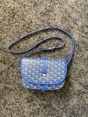 Blå axelväska  - Snygg blå axelväska med vitt grafiskt mönster och justerbar axelrem. Väskan har ett lock med spänne framtill och rundade kanter. Materialet ser ut att vara tåligt canvas med detaljer i konstläder. Perfekt för dig som vill ha en trendig och unik accessoar.