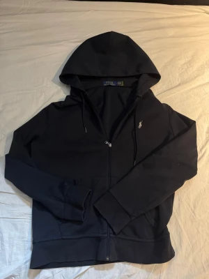 Svart hoodie från Polo Ralph Lauren - Säljer en mörkblå hoodie från Polo Ralph Lauren med dragkedja framtill och klassisk huva. Tröjan har fickor på magen och den ikoniska broderade loggan i vitt på bröstet. Perfekt för en avslappnad och stilren look. Köpt för 1950kr i Palma de mallorca i en ralph lauren butik för ca 1 år sedan. Den är äkta och är i storlek M.