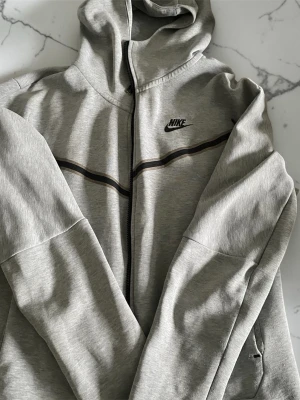 Grå zip hoodie från Nike - Snygg grå hoodie från Nike med dragkedja och svart logga på bröstet. Hoodien har en tunn svart linje över bröstet och huva. Tillverkad i mjukt material som känns skönt mot huden. Perfekt för en avslappnad och sportig stil.