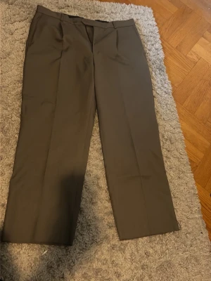Mörkbruna raka kostymbyxor - Snygga mörkbruna kostymbyxor med rak passform och pressveck. Byxorna har hällor för skärp, två sidofickor fram och två bakfickor med knapp. Materialet är slät och lite glansig väv, perfekt för en stilren look.