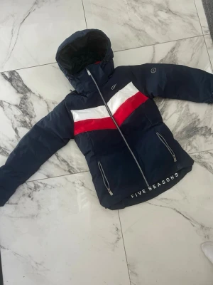 Marinblå skidjacka från Five Seasons - Snygg marinblå vindjacka med huva från Five Seasons. Jackan har vita och röda detaljer över bröstet, två dragkedjeförsedda fickor och logga på nederkanten. Perfekt för blåsiga dagar och sportig stil. Använd en skidsemester så fint skick!stl S