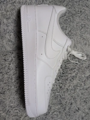 White Nike Air Force 1 sneakers - Klassiska helvita Nike Air Force 1 sneakers i lågt utförande. Skorna har en stilren design med vit sula, vita skosnören och den ikoniska swoosh-loggan på sidan. Tillverkade i slätt läder med perforeringar på tån för extra ventilation. Aldrig använt dem har bara legat i skohyllan.