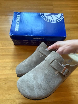 Birkenstock Boston taupe mockatofflor - Säljer ett par snygga beige/ljusgrå Birkenstock Boston i storlek 42. Som nya och i mycket fint skick utan några brister.  Klassisk och bekväm modell i mjuk mocka med justerbart metallspänne och ergonomisk fotbädd i kork. Perfekta både hemma och ute!  Originalkartong följer med.  Vid bud eller funderingar är det bara att skriva! 😊