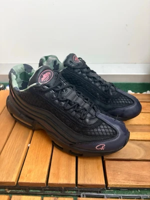 Svarta sneakers med camo-foder - Säljer ett par svarta sneakers med mesh och läderdetaljer, rosa logga på plösen och sidan samt camo-mönstrat foder. Snygg chunky sula och coola detaljer som sticker ut. Perfekta för dig som vill ha något unikt till din streetwear-look.
