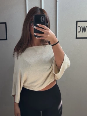 Ljusbeige offshoulder topp Nelly - Trendig ljusbeige offshoulder topp med lös passform och trekvartsärm. Toppen har en avslappnad och croppad stil som passar perfekt till höga byxor eller kjol. Materialet känns mjukt och luftigt, vilket gör den skön att bära under varmare dagar.