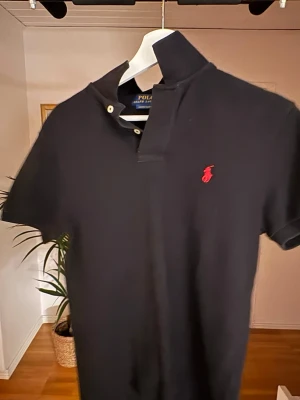 Svart pikétröja från Polo Ralph Lauren - Klassisk svart pikétröja från Polo Ralph Lauren med röd broderad logga på bröstet. Tröjan har korta ärmar, krage och knappar vid halsen. Modellen är custom slim fit och passar perfekt till en clean och stilren look.
