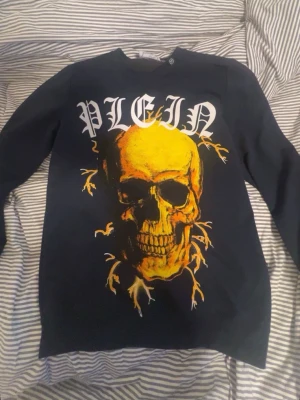 Svart långärmad t-shirt Philipp Plein - Svart långärmad t-shirt från Philipp Plein med stor gul dödskalle och vit text 'PLEIN' på bröstet. Snygg och edgy design med detaljer i gult och vitt. Perfekt för dig som vill sticka ut med en unik streetwear-look.