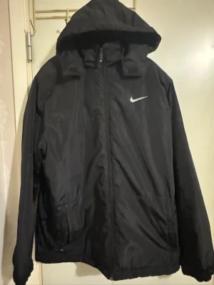 Svart Nike vindjacka med luva  - Svart vindjacka från Nike med luva och dragkedja framtill. Jackan har två sidofickor och en vit Nike-logga på bröstet. Perfekt för blåsiga dagar och enkel att matcha med olika outfits. Materialet är lätt och vattenavvisande.