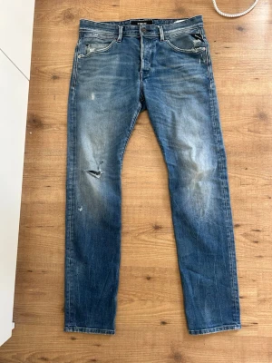 Blå slitna jeans från Replay - Säljer ett par blå jeans från Replay med slitningar och lätt tvättad look. Modellen har klassisk femficksdesign, dragkedjegylf och raka ben. Perfekta för en avslappnad och trendig stil.