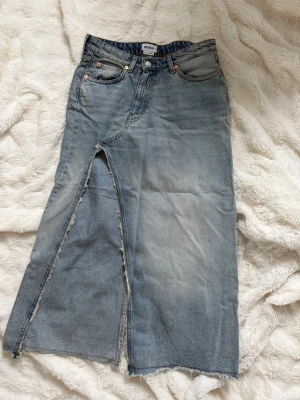 Lång jeanskjol med slits - Säljer en lång jeanskjol i ljusblå denim med råa kanter och en cool slits framtill. Kjolen har klassiska fem fickor och knappgylf. Perfekt för dig som gillar en avslappnad och trendig stil.
