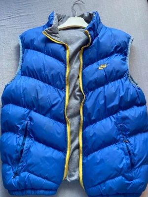 Blå och grå vändbar dunväst Nike - Snygg vändbar dunväst från Nike i blått och grått med gul dragkedja. Ena sidan är blå med gul Nike-logga, den andra är ljusgrå med svart logga. Västen har ståkrage och fickor med dragkedja. Perfekt för lager-på-lager och streetwear. Inte äkta 