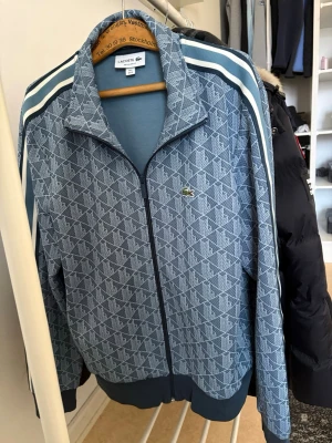 Blå mönstrad ziptröja från Lacoste - Säljer en blå ziptröja från Lacoste med vitt geometriskt mönster och klassiska ränder på axlarna. Tröjan har dragkedja framtill, ståkrage och ribbade muddar. Ikoniska krokodilloggan på bröstet köpt för 2lax i butik 