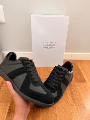Maison Margiela GATs - Stilrena svarta sneakers från Maison Margiela med en mix av mocka och slätt läder. Skorna har svarta snören, diskreta paneler och en klassisk låg siluett. Sulan är i gummi med unikt mönster och logotyp under. Perfekta för dig som gillar minimalistisk design.