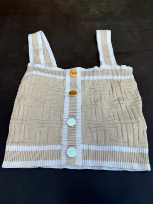 Beige ribbad linnetopp från Amisu - Trendig beige och vit ribbad linnetopp från Amisu med breda axelband och dekorativa guldknappar framtill. Toppen har ett diskret geometriskt mönster i tyget och är croppad i modellen. Perfekt för en cool och avslappnad stil.