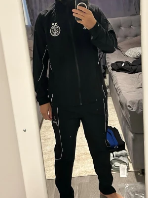 Svart PSG träningsset från Jordan - Säljer ett svart träningsset från Jordan x PSG med vita detaljer. Setet består av en jacka med dragkedja och matchande byxor. PSG-logga och Jordan-logga på bröst och ben. Perfekt för träning eller chill. Materialet är lätt och andas.