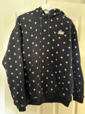 Svart hoodie med vita stjärnor SDMN - Snygg svart hoodie från SDMN med vita stjärnor över hela plagget. Hoodien har en stor känguruficka framtill, justerbar huva med snören och ribbade muddar. Perfekt för dig som gillar streetwear och vill sticka ut lite extra.