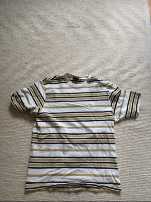 Stussy Randig t-shirt i vitt och gult - Säljer en fräsch t-shirt med vita, gula och svarta ränder. T-shirten har klassisk passform och korta ärmar. Perfekt för dig som gillar en enkel men ändå stilren look. Materialet känns mjukt och skönt mot huden.