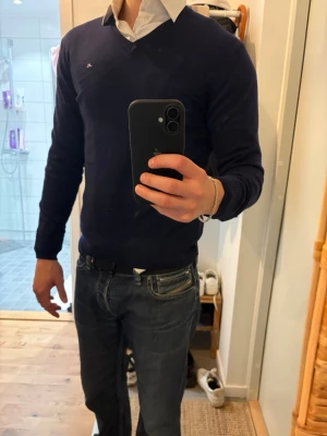 Mörkblå V-ringad tröja herr J-lindeberg - Snygg mörkblå V-ringad tröja med diskret logga på bröstet. Tröjan har en klassisk passform och långa ärmar, perfekt att bära över en skjorta för en clean och stilren look. Passar dig som gillar enkel och tidlös stil.