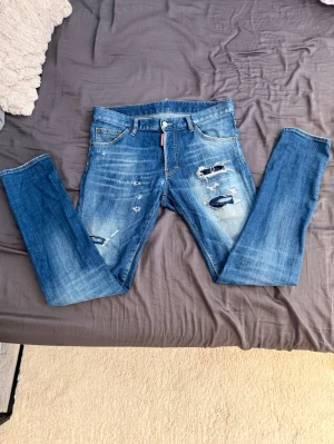 Blåa dsquered2 jeans - Säljer ett par blå jeans med slitningar och hål på framsidan. Jeansen har en klassisk femficksmodell, normal midja och raka ben. Snyggt tvättade detaljer och råa kanter ger en trendig look. Perfekta för dig som gillar en avslappnad och edgy stil. Jeansen är från märket dsquered2 och dom är äkta, dom är i passformen slim