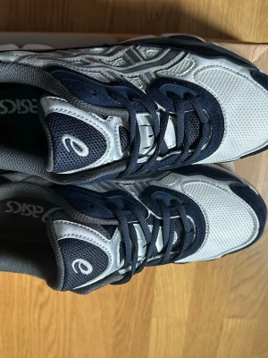 Asics vita och marinblå sneakers - Snygga sneakers från Asics i vitt och marinblått med mesh- och syntetpaneler. Skorna har en sportig design med tydliga Asics-loggor på sidan och tungan. Yttersulan är robust och mönstrad för bra grepp. Perfekta för dig som gillar en retroinspirerad look.