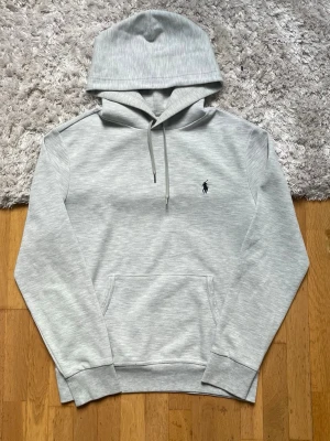 Ljusgrå hoodie från Polo Ralph Lauren - Snygg ljusgrå hoodie från Polo Ralph Lauren med klassisk huva och dragsnören. Tröjan har en broderad logga på bröstet och en stor magficka. Tillverkad i mjukt bomullsmaterial och har ribbade muddar vid ärmslut och nederkant. Ena metall saken på ett snöre har åkt av men märks inte vid användning. Annars är hoodien i galant skick