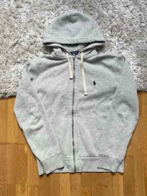 Grå zip hoodie från Polo Ralph Lauren - Snygg ljusgrå hoodie med dragkedja från Polo Ralph Lauren. Klassisk design med huva och vita dragsnören, samt den ikoniska mörkblå loggan broderad på bröstet. Tillverkad i mjukt sweatshirtmaterial och har fickor framtill.