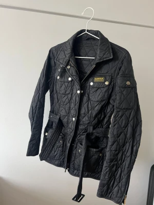 Svart quiltad jacka från Barbour storlek 32 - Snygg svart quiltad jacka från Barbour med bälte i midjan, gulddetaljer och flera fickor. Jackan har dragkedja och tryckknappar framtill samt Barbour International-logga på bröstet. Perfekt för höst och vår.