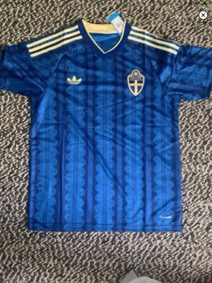 Blå Sverige fotbollströja Adidas - Säljer en blå fotbollströja från Adidas med Sveriges landslagsemblem på bröstet. Tröjan har gula ränder på axlarna, mönstrad design och korta ärmar. Perfekt för dig som vill stötta Sverige med stil på matchen.