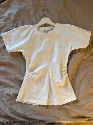 Snygg T-shirt från only - Säljer en vit t-shirt i bomull med figurnära passform och rynkade detaljer vid midjan. T-shirten har rund halsringning och korta ärmar. Perfekt för dig som gillar en enkel men ändå unik look.