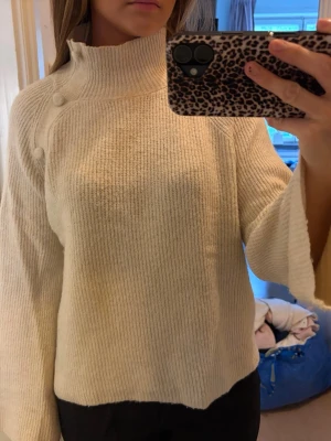 Krämvit stickad polotröja med knappar - Säljer en krämvit stickad tröja med hög polokrage och dekorativa knappar på axeln. Tröjan har vida ärmar och är ribbstickad i ett mjukt material. Perfekt för dig som gillar en clean och stilren look, den har dock en smink fläk längst fram på kragen.