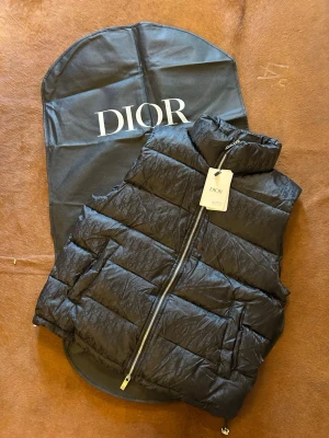 Svart dunväst från Dior - Snygg svart dunväst från Dior med diskret monogrammönster i tyget. Västen har hög krage, dragkedja framtill och två stora fickor. Perfekt för lager-på-lager och ger en exklusiv vibe till din outfit.