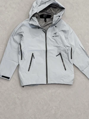 Arc'teryx regnjacka - Snygg ljusblå regnjacka från Arc'teryx med huva och hel dragkedja framtill. Jackan har två snedställda fickor med dragkedja, justerbara ärmslut med kardborre och vattentätt material. Perfekt för regniga dagar och outdoor-äventyr.