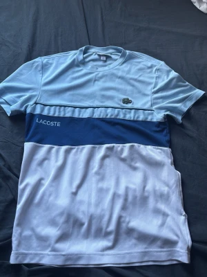 Blåvit t-shirt från Lacoste - Snygg t-shirt från Lacoste i ljusblått, mörkblått och vitt. Klassisk krokodil-logga på bröstet och Lacoste-tryck på sidan. T-shirten har korta ärmar och är gjord i mjuk bomull, perfekt för en avslappnad stil. Har ett litet hål u sidan på tröjan men inget mörk värdigt 