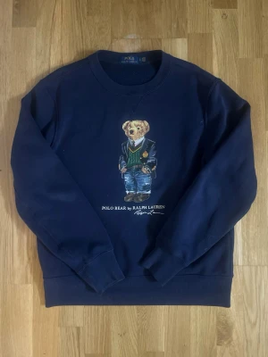 Blå sweatshirt Polo Bear Ralph Lauren - Mörkblå sweatshirt från Ralph Lauren med den ikoniska Polo Bear-trycket på bröstet. Rund halsringning, långa ärmar och ribbade muddar. Tillverkad i mjukt bomullsmaterial för en skön och avslappnad look. Perfekt för dig som gillar klassisk streetwear med en twist. Skriv gärna om ni har frågor mm, priset kan diskuteras vid rimliga bud.