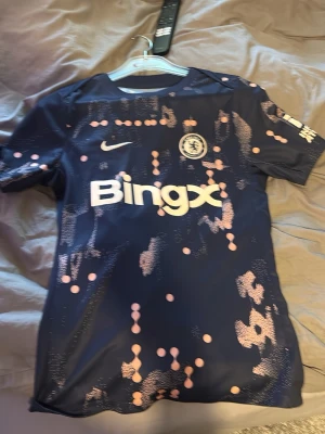 Chelsea FC mörkblå matchtröja Nike - Säljer en Chelsea FC matchtröja i mörkblått med rosa och vita detaljer. Tröjan har Bingx-logga på bröstet, Nike-logga och Chelsea-emblem. Modern design med korta ärmar och rund hals, perfekt för fotboll eller träning.