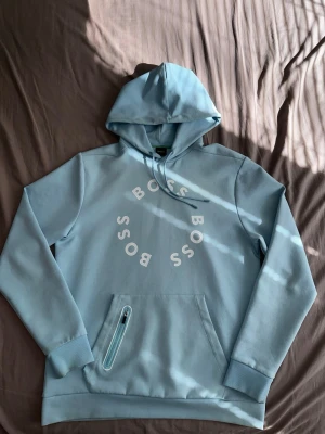  BOSS hoodie - Snygg ljusblå hoodie med huva och dragsko. Stort cirkelformat BOSS-tryck på bröstet i vitt. Praktisk magficka med dragkedjedetalj. Tillverkad i mjukt material, perfekt för en avslappnad stil.