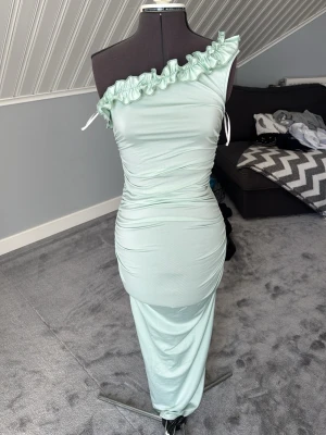 Ljusgrön oneshoulder långklänning - Snygg ljusgrön långklänning med oneshoulder-design och volangdetalj längs axeln. Klänningen har en figurnära passform och är tillverkad i ett mjukt, stretchigt material. Perfekt för dig som vill sticka ut med en unik och trendig look.