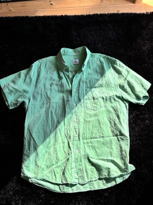 Fin grön kortärmad skjorta från Lacoste - Säljer en grön kortärmad skjorta från Lacoste i linne. Skjortan har klassisk krage, knappstängning framtill och en bröstficka med den ikoniska krokodilloggan. Perfekt för varma dagar och enkel att styla till olika outfits.