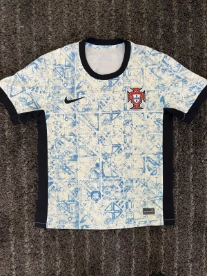 Portugal Nike fotbollströja blåvit - Portugal fotbollströja från Nike i vitt med ljusblått mönster och svarta detaljer. Tröjan har korta ärmar, svart krage och sidopaneler. Portugal-emblemet sitter på bröstet tillsammans med Nike-loggan. 