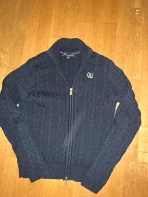 Mörkblå kabelstickad kofta med dragkedja - Mörkblå kabelstickad kofta från JR Collection med dragkedja framtill och hög krage. Koftan har ett broderat emblem på bröstet och ribbade muddar vid ärmslut och nederkant. Perfekt för lager på lager och en klassisk look.