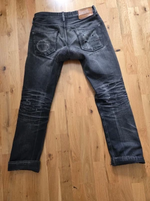 Indigofera selvedge jeans - Säljer ett par svarta jeans med raka ben och slitningar på både fram- och baksida. Jeansen har klassiska fem fickor, tydliga whiskers och en patch bak i midjan. Materialet är denim i bomull och passformen är relaxed. Inga hål men flertalet lagningar samt trasiga fickor.