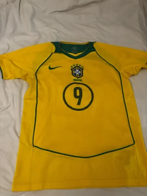 Gul Brasil Ronaldo 9 fotbollströja - Säljer en klassisk gul Brasilien fotbollströja från Nike med gröna detaljer. Tröjan har nummer 9 och Ronaldo tryckt på ryggen samt Brasiliens landslagsemblem på bröstet. Perfekt för dig som älskar fotboll och vill hylla en legend.